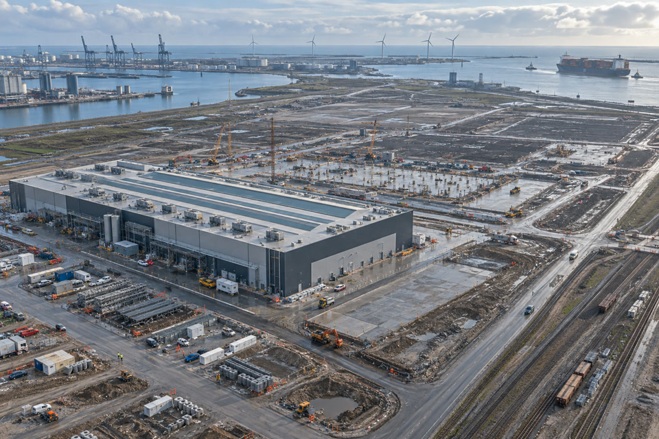 Vue aérienne synthétique d’un chantier industriel côtier à Dunkerque, avec une première halle achevée, de vastes fondations, le port et des éoliennes.